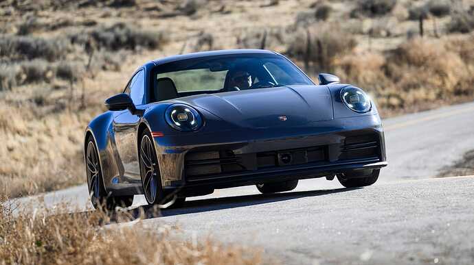 front view of a dark colored sports car driving on a winding road surrounded by dry grass and hills.